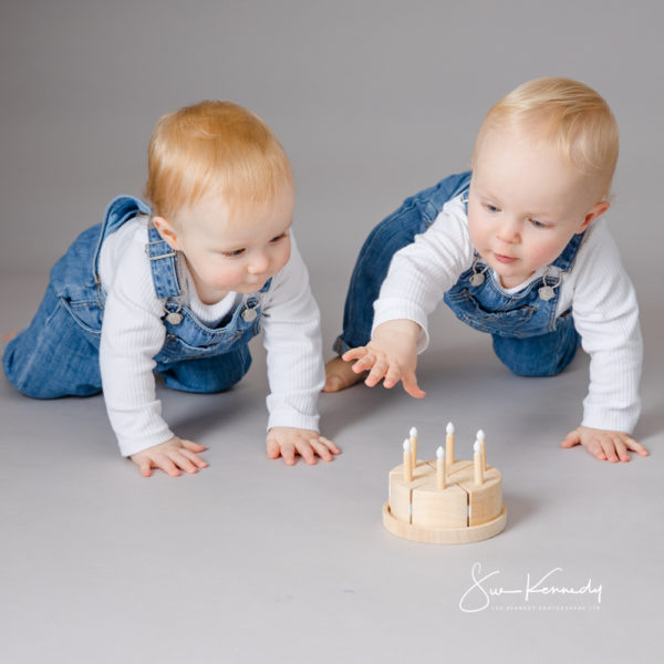 Two blonde baby twins in denim dungarees and white tops crawling towards a small wooden birthday cake prop during their first birthday portrait session.