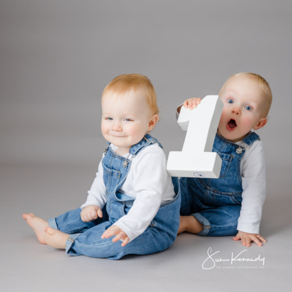 Twin babies in denim dungarees during their first birthday session, one smiling quietly while the other excitedly holds a large wooden number one.