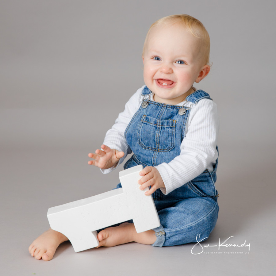 Smiling baby boy in denim dungarees holding a wooden number one during his first birthday portrait session.