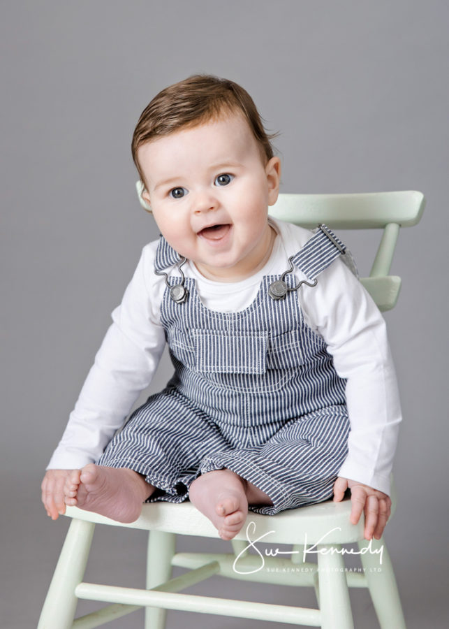 Smiling six-month-old baby boy sitting on a cream Ercol child's chair wearing striped dungarees and a white top - studio portrait by Sue Kennedy Photography, specialist baby photographer in Harlow, Essex.