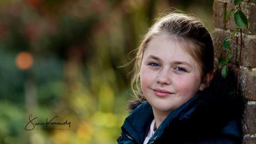 10 year old girl leaning against a wall with blurry background on greenery behind