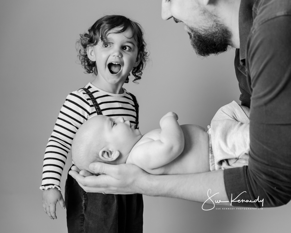 big sister expressing surprise at being asked to kiss baby brother held by their father at a recent photography session
