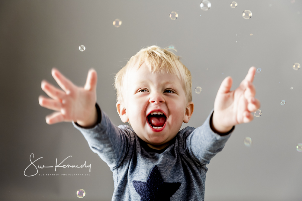 Toddler boy playing with bubbles with his arms outstretched to catch some.