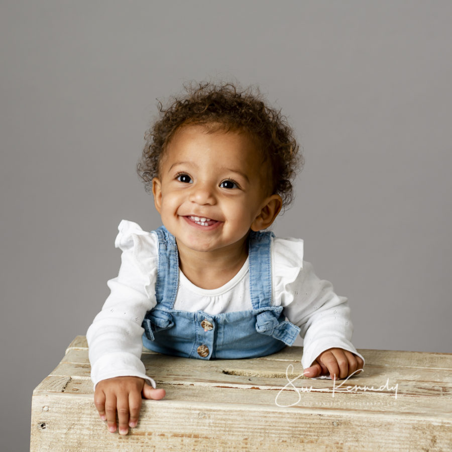 baby smiling at the camera in an outfit of white t-shirt and denim dungarees