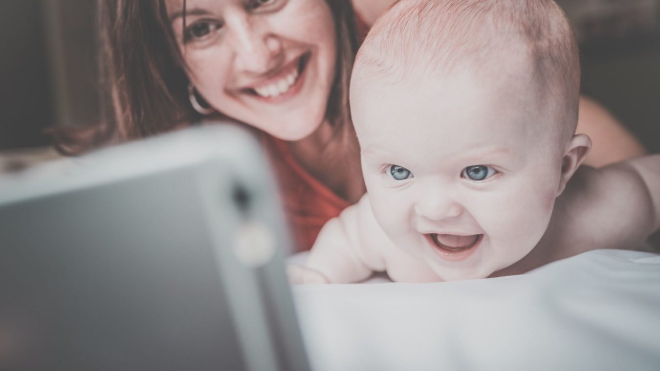 Baby smiling at a mobile phone with Mum in the background