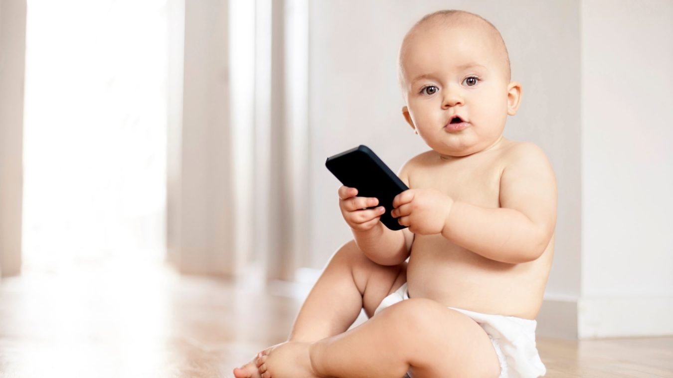 Baby sat on the floor in a nappy looking toward camera, and holding a smartphone