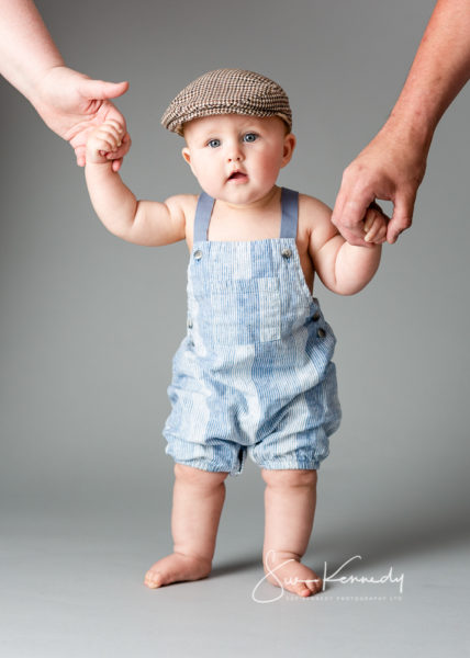 Toddler boy dressed in dungarees and a flat cap standing holding parents hands.