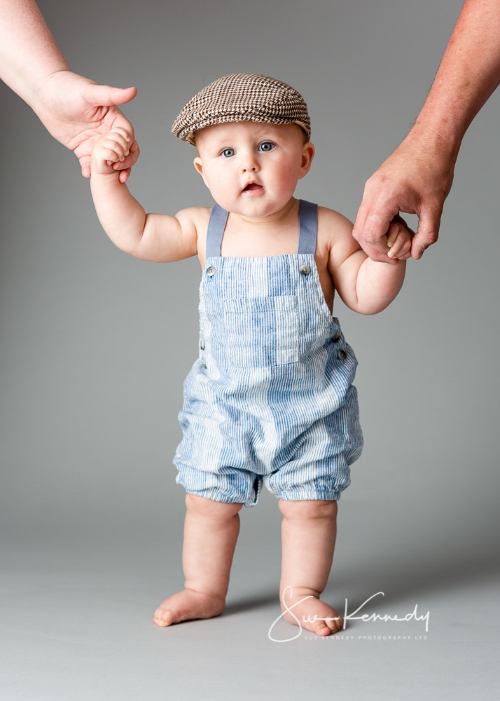 Toddler boy dressed in dungarees and a flat cap standing holding parents hands