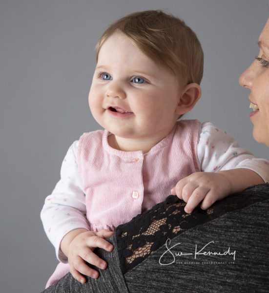 A baby girl from Harlow in Essex is being held by her Mum and smiling over her shoulder to Dad standing off-camera during her second of three photoshoots to mark the baby's first year.