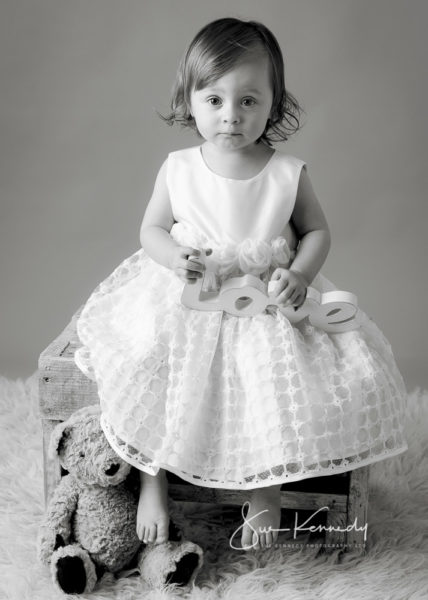 Toddler girl from Harlow in Essex sat on a stool looking toward camera holding a wooden love sign, presented as a black & white portrait.