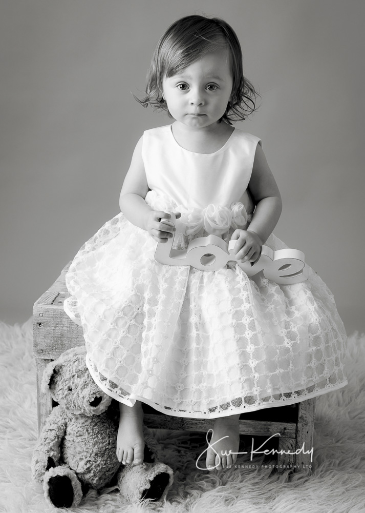 Black and white portrait Photographs of a toddler girl sat on a stool with a teddy at her feet in her party dress.