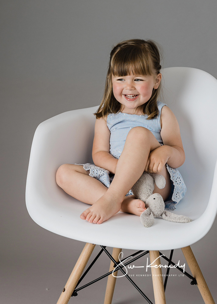Little girl sat casually on a modern white chair smiling at camera during a professional kids portrait session in Harlow, Essex