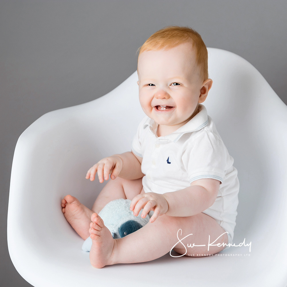 Baby boy sat on a modern white chair laughing. Photography by Sue Kennedy, Harlow, Essex