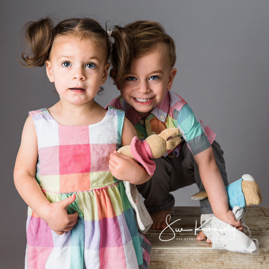 Twin boy and girl standing together, with the little boy grinning slightly behind his sister and both holding their soft toy rabbits. Photography by Sue Kennedy, Harlow, Essex