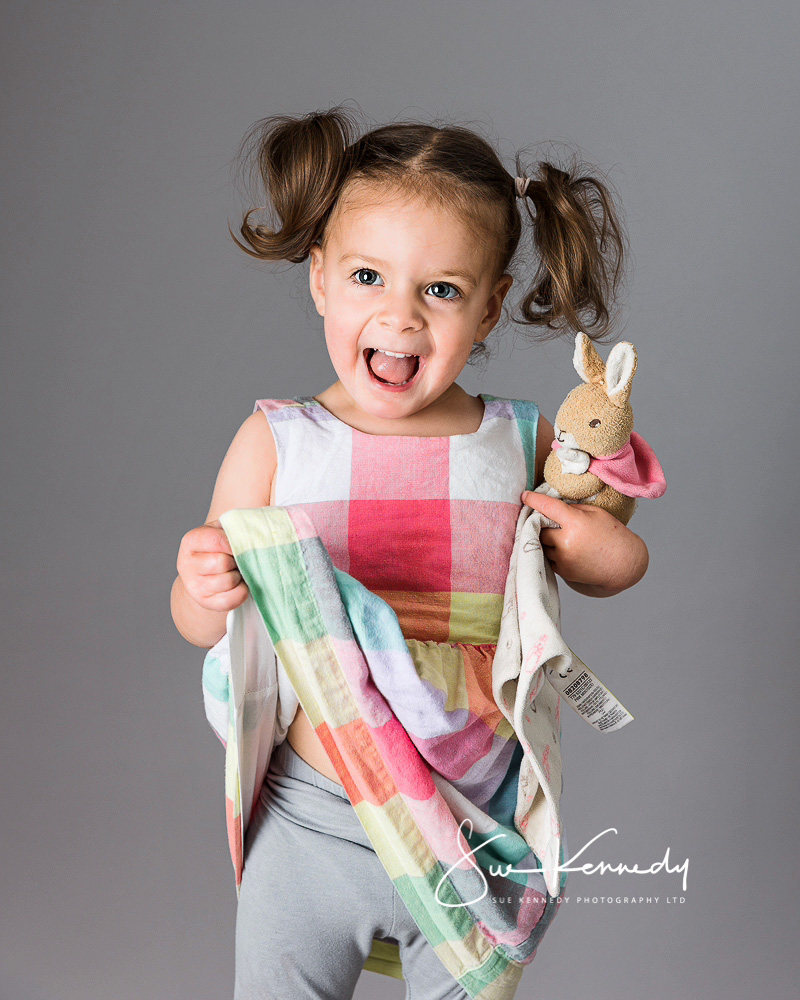 Toddler girls standing and lifting her dress up with an excited expression on her face, whilst holding her bunny toy. Photography by Sue Kennedy Photography, Harlow, Essex