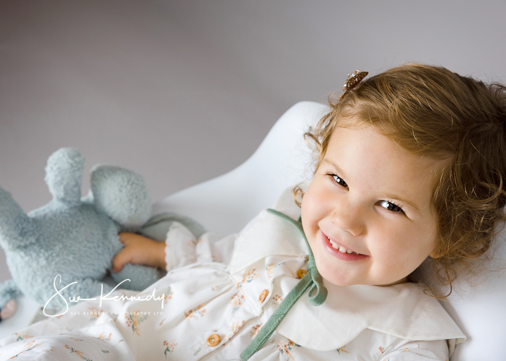Toddler girl lying on a chair smiling to camera holding her favourite toy