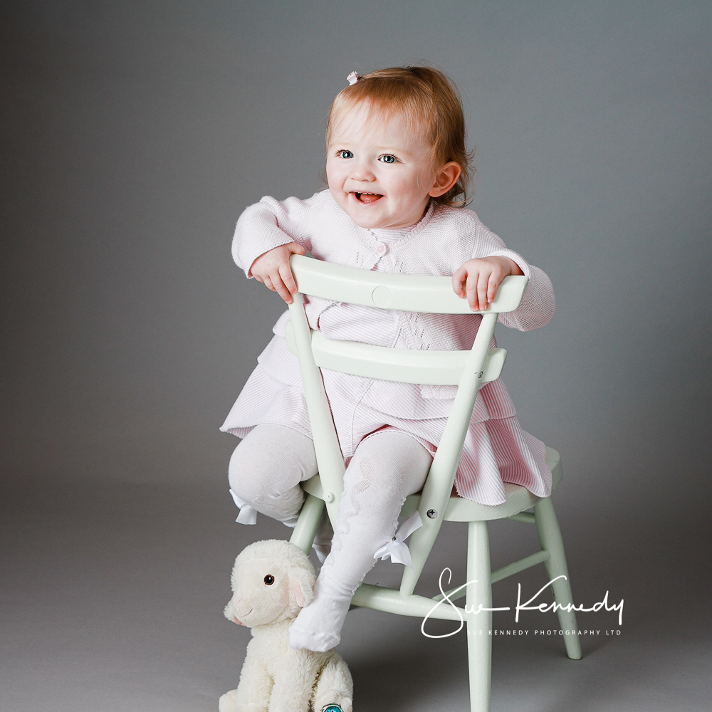 Toddler girl sat on a white children's chair with her lammy soft toy by her feet.