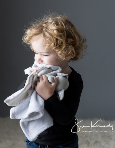 Toddler boy cuddling his two elephant comforters looking away from camera toward the llight