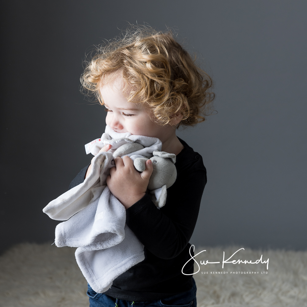 Toddler boy cuddling his two elephant comforters looking away from camera toward the llight