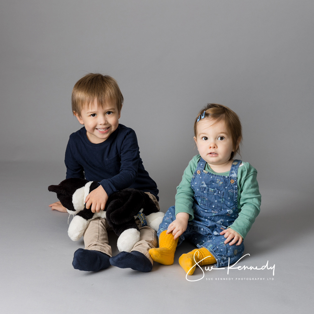 Brother and sister sitting on a grey backdrop with Jessie the cat soft toy between them.