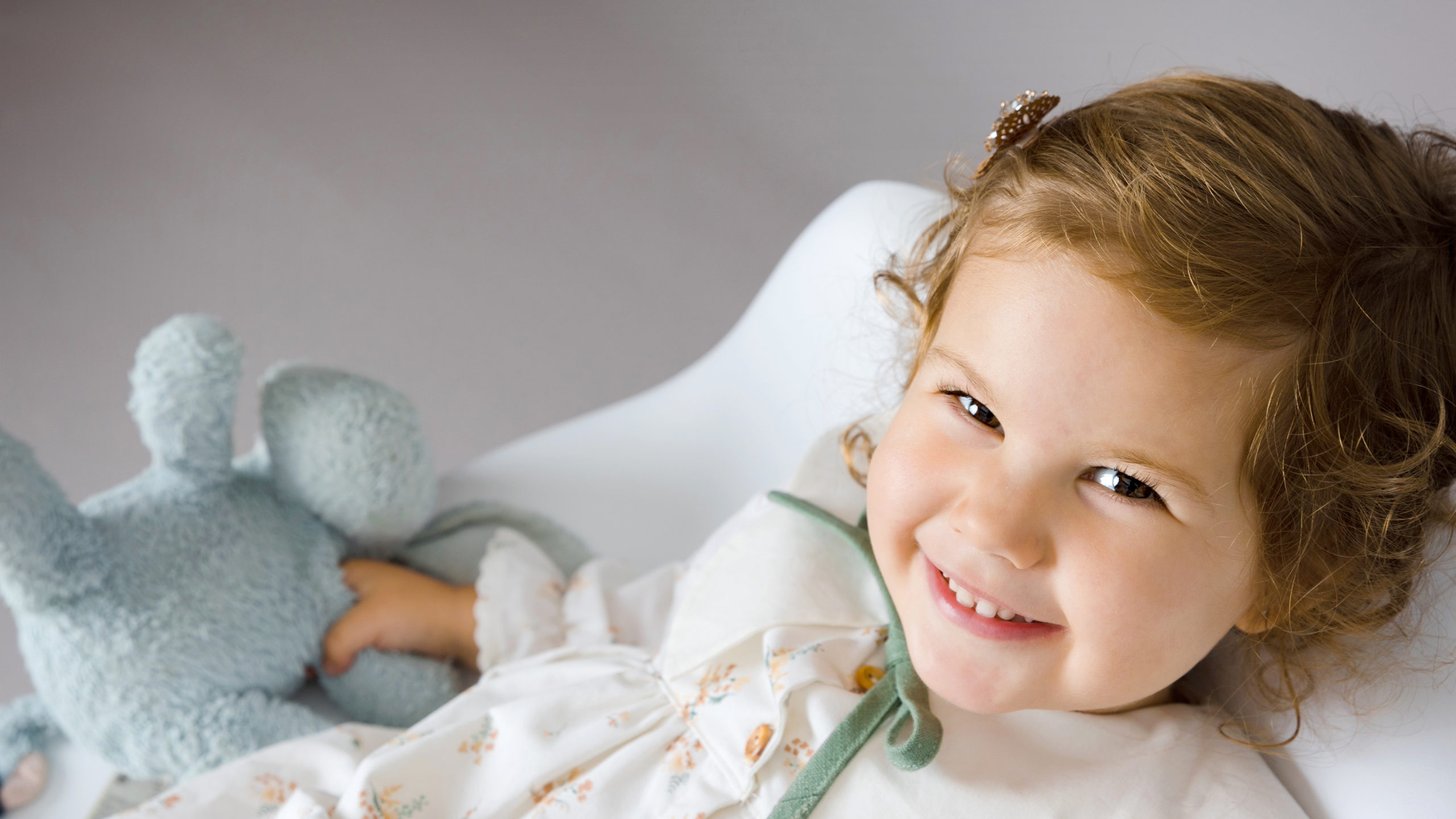 Child phootgraphed from above lying back on a white chair holding a soft toy in her hand.