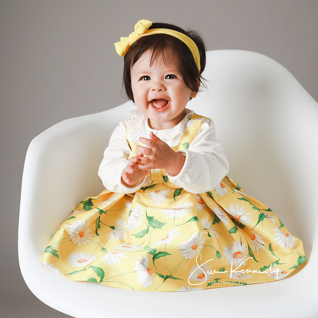 Baby girl sat on a contemporary white chair in a yellow dress clasping her hands together and smiling at the photographer.