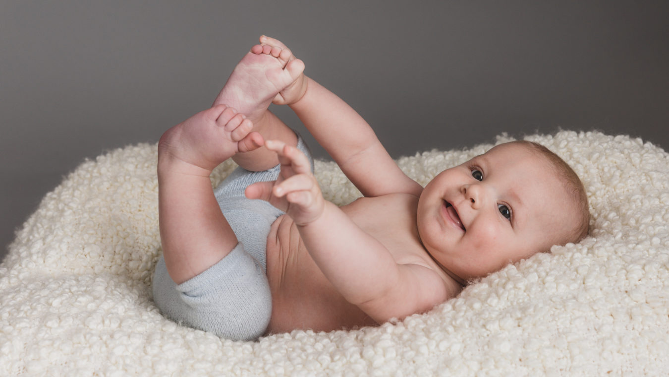 Professional baby portrait – 5-month-old baby lying on his back, holding his feet and smiling at the camera, taken at Sue Kennedy Photography studio in Harlow, Essex.