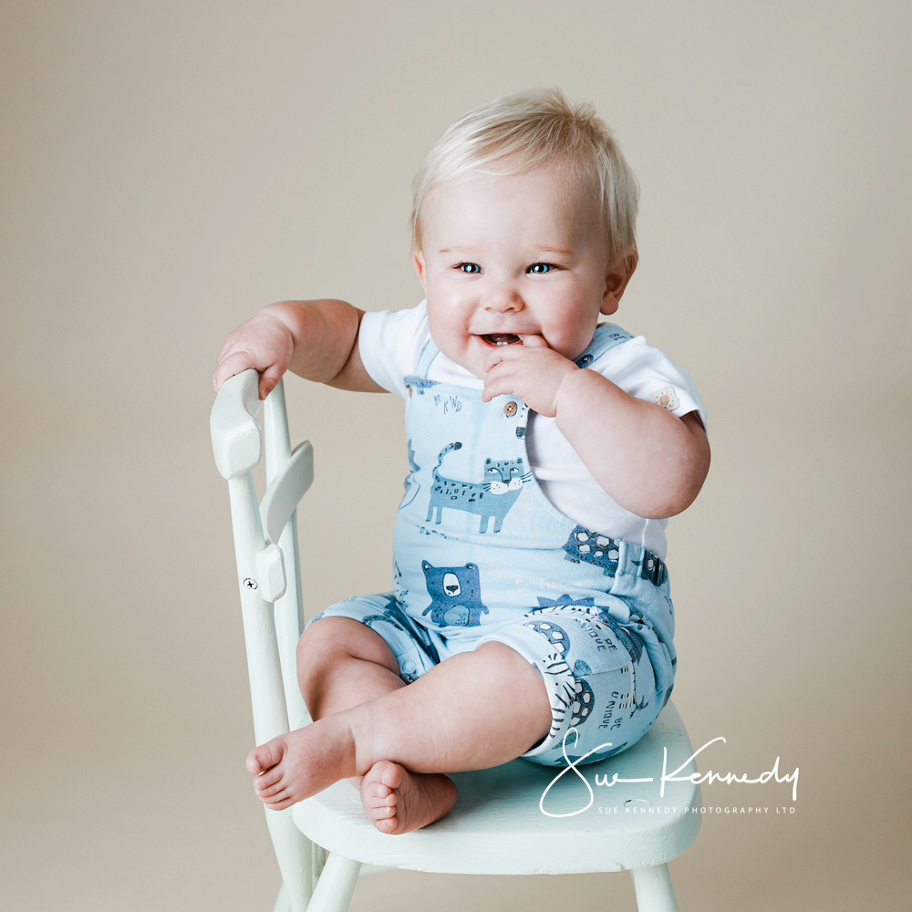 12 month old baby sat on a small child's size chair and smiling to camera