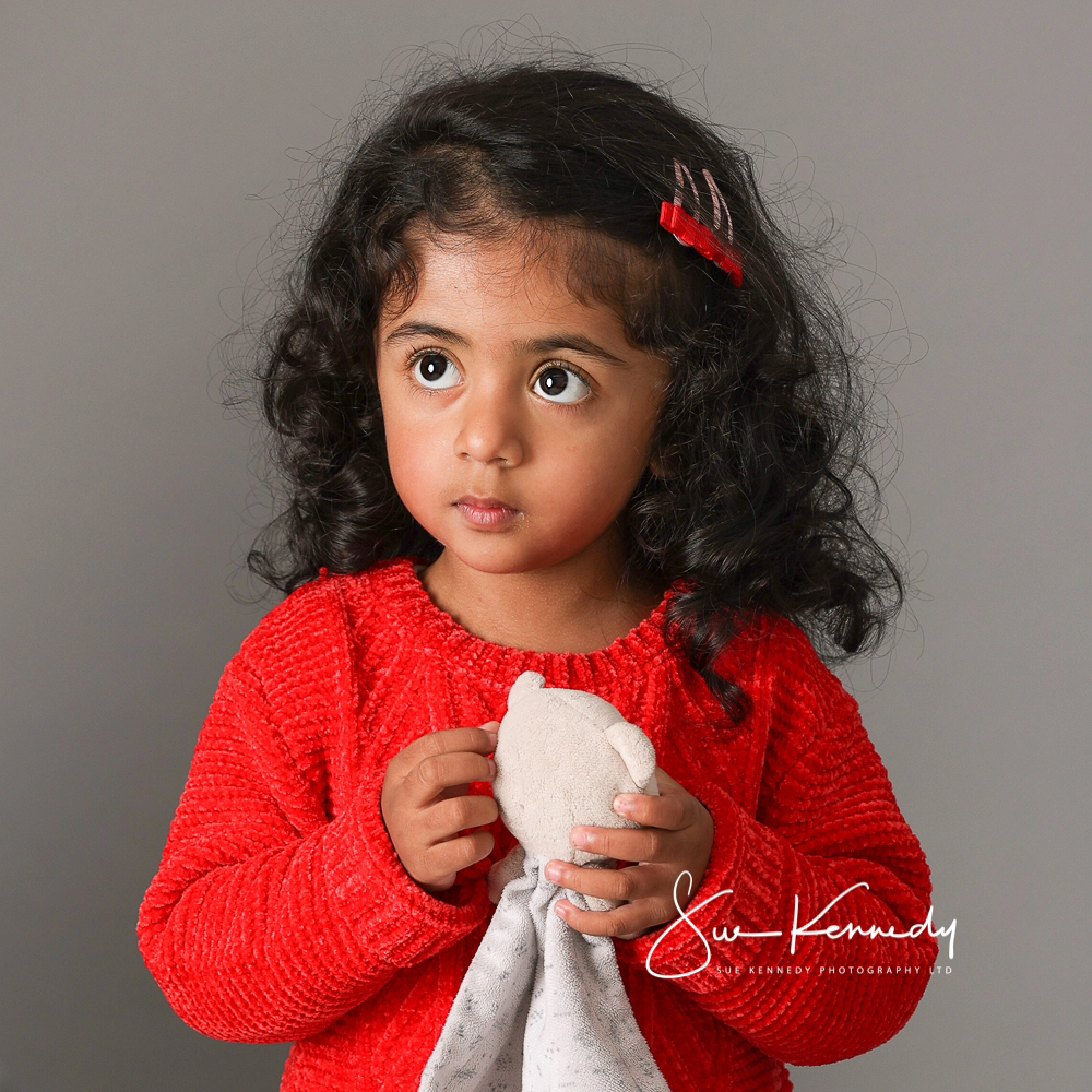 Young girl holding her soft toy comforter and looking away from camera, wearing a red jumper.