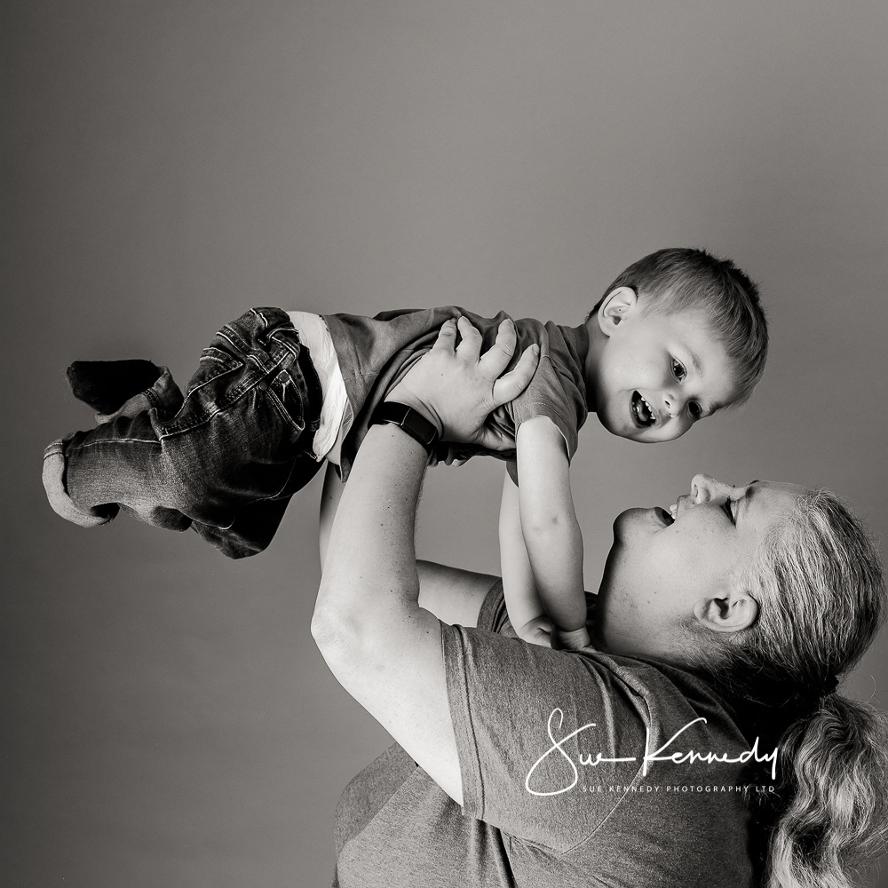 Toddler boy being held aloft by his mummy, both smiling. Black and white photograph showing the love and connection between them.