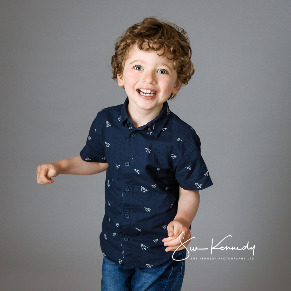Three year old boy standing and smiling towards camera against a grey background dressed in a navy blue shirt and jeans