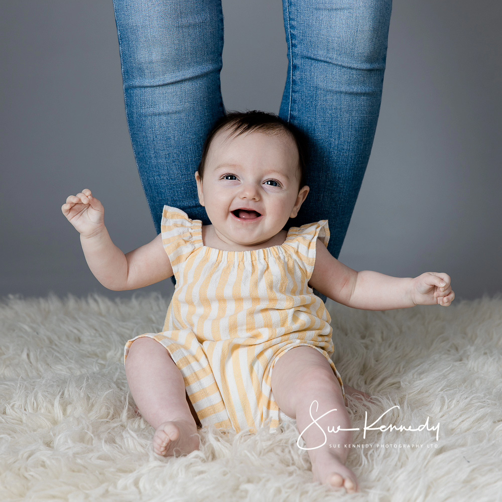 Baby sitting at mums feet for support on a fluffy cream blanket, throwing her arms around smiling