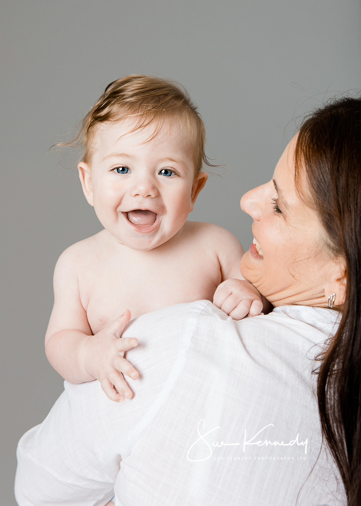 Baby boy smiling at camera looking over his mothers shoulder as she looks lovingly at him