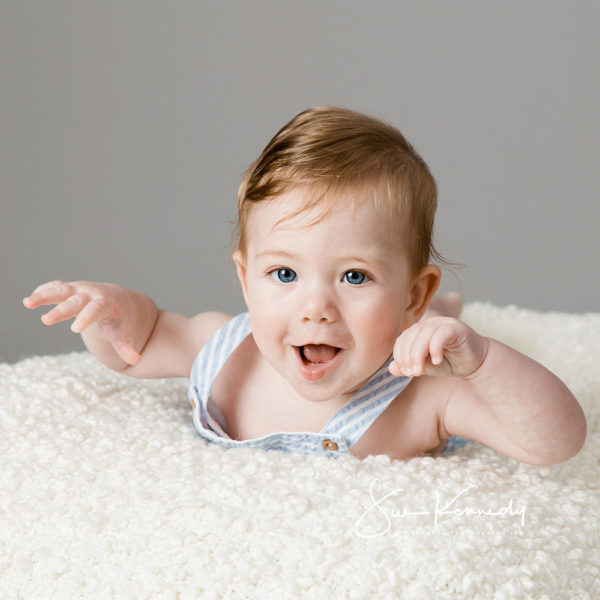 6 month old baby lying on his tummy on a cream blanket looking excited and lifting his arms in the air during a relaxed baby portrait session at Sue Kennedy Photography in Harlow, Essex.