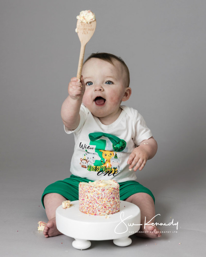 Baby lifting a wooden spoon covered in cake during a first-birthday cake smash photographic experience, smiling with a sprinkle-covered cake in front of them.