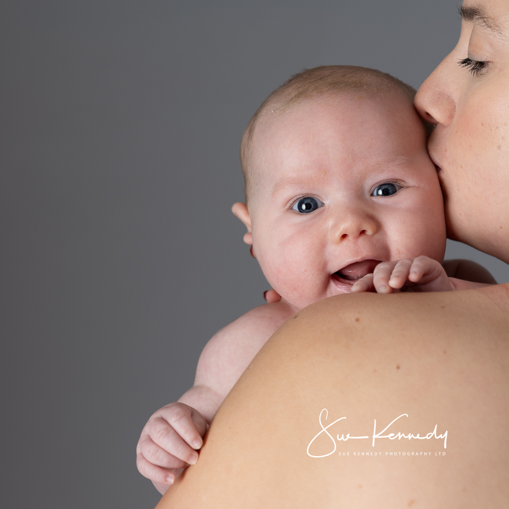 A baby with bright eyes and a joyful expression is held close on his mother's shoulder, receiving a gentle kiss. Photographed in studio by Sue Kennedy Photography.