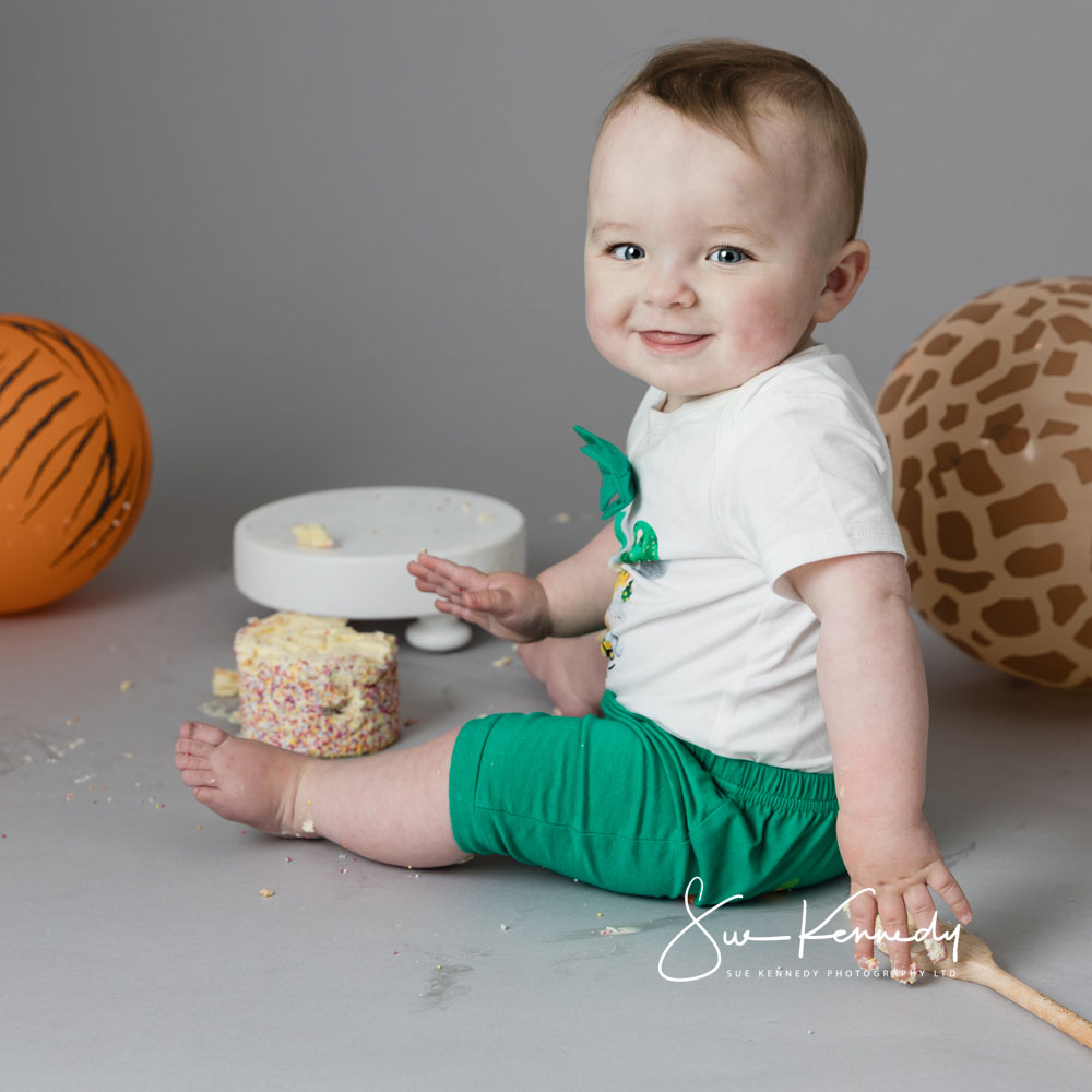 Smiling baby boy in green shorts during a first birthday portrait session, sitting near a half-smashed sprinkle cake with animal-print balloons in the background – photographed by Sue Kennedy Photography.