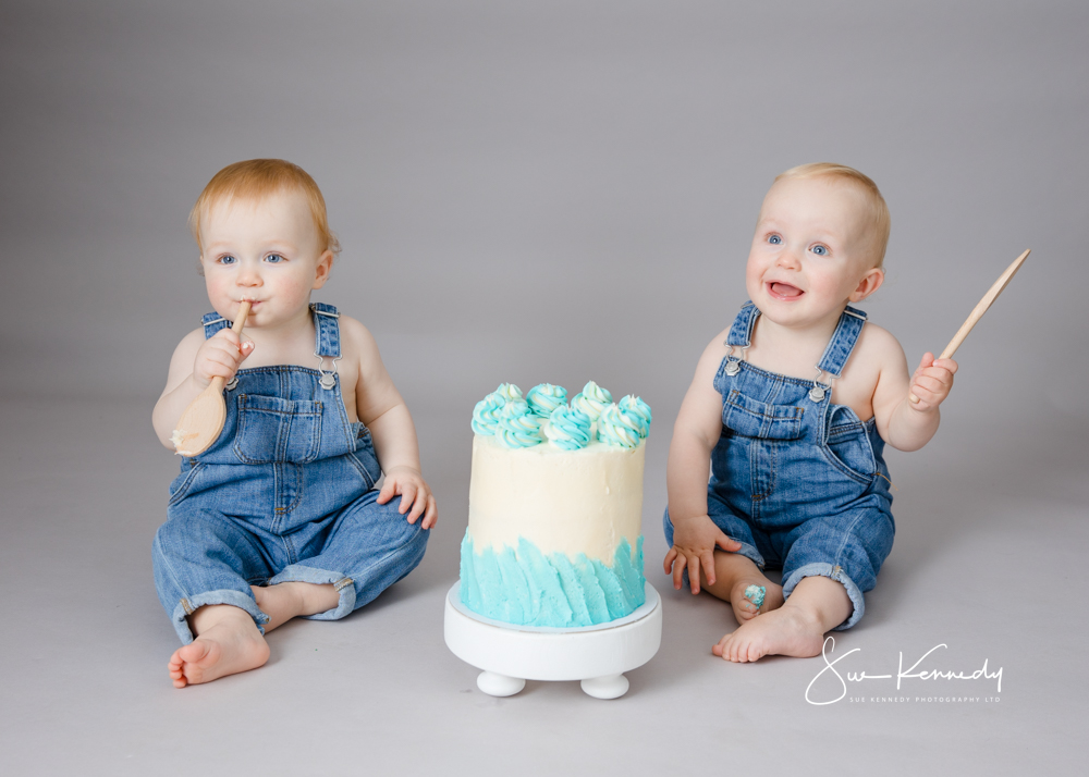 Twin babies in denim dungarees enjoying a blue and white first birthday cake smash in Sue Kennedy’s Harlow studio, each holding a wooden spoon against a soft grey background.