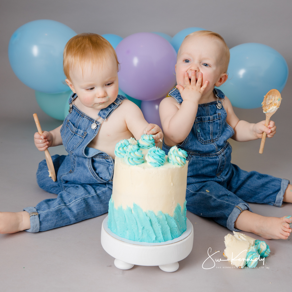 Twin babies in denim dungarees sharing a blue-and-white birthday cake during a cake smash at Sue Kennedy’s Harlow studio, with pastel balloons in the background.