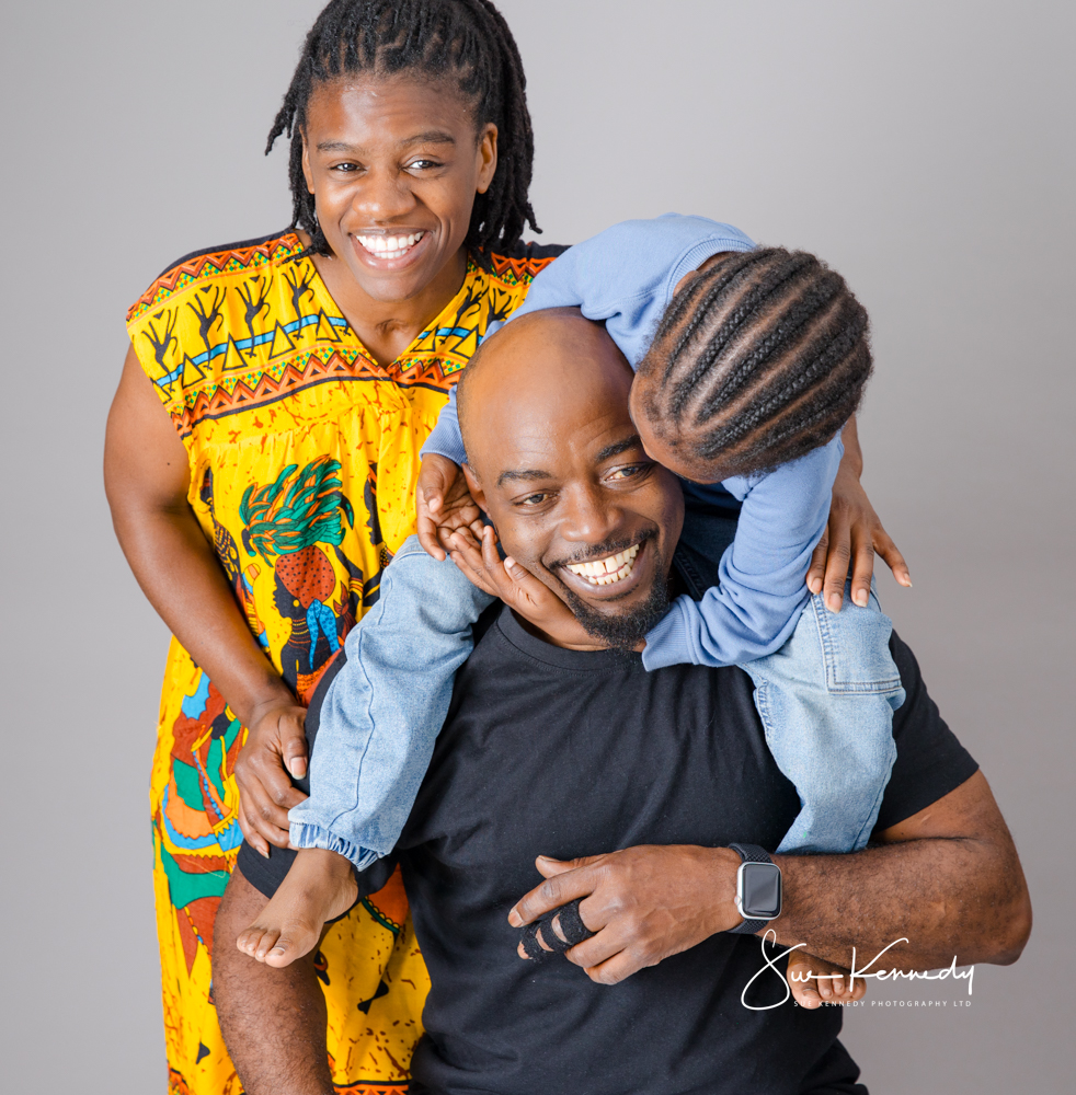 A young child playfully climbing onto a parent’s shoulders while another parent stands beside them smiling in a studio setting.