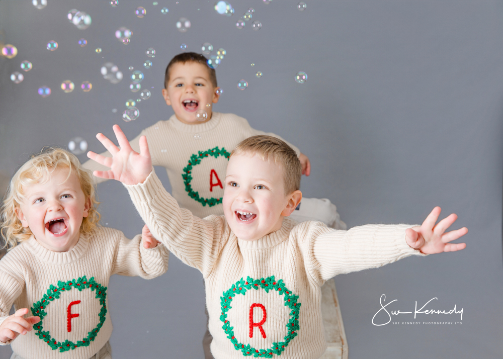 Three young children wearing cream festive jumpers playing with bubbles in the studio, smiling and reaching with excitement.