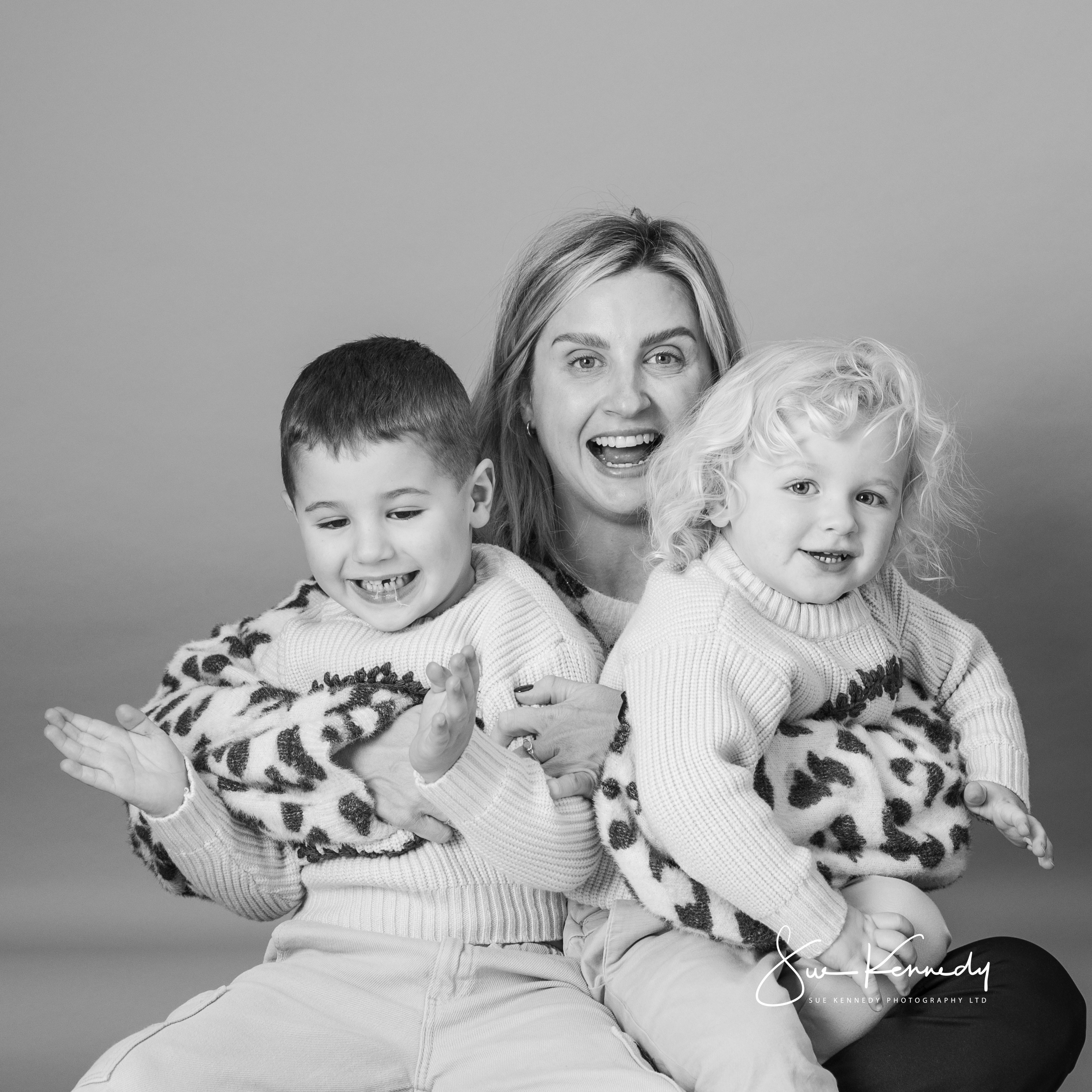 Black-and-white studio portrait of a mother with her two young children, capturing their playful energy and close bond.