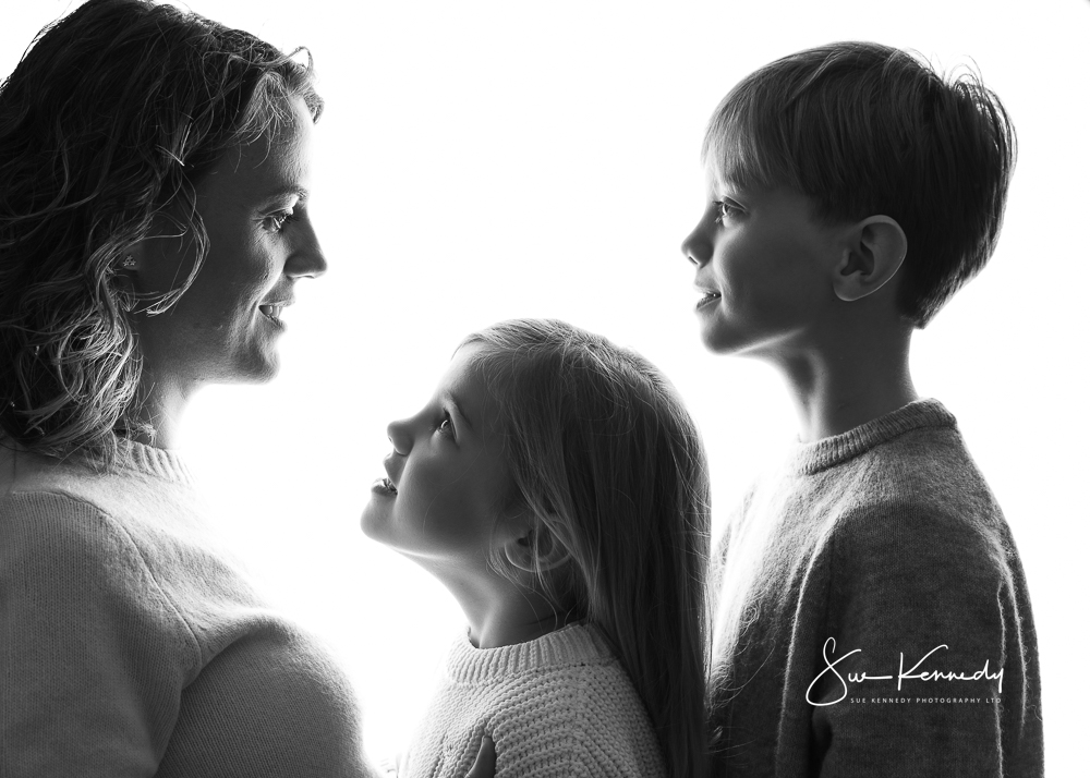 Mother and her two children in a black-and-white studio portrait, facing each other with soft expressions and natural connection.