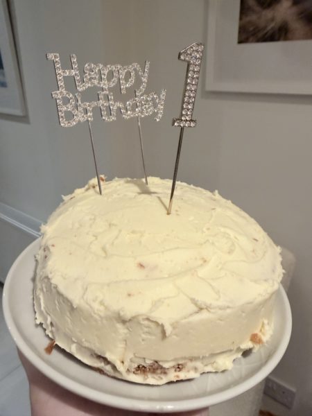 A homemade first birthday cake smash cake on a white cake stand, generously covered in white buttercream frosting with a rustic, hand-spread finish. Two sparkly rhinestone cake toppers are pushed into the top - one reading 'Happy Birthday' and a number '1'.